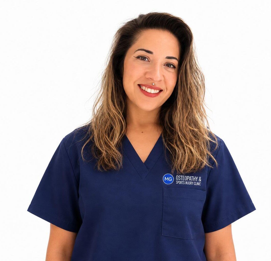 Friendly woman in navy scrubs smiling at the camera, logo on chest reads 'MG Osteopathy & Sports Injury Clinic'